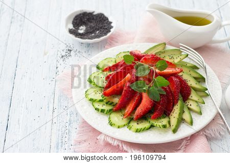Vegan salad from strawberry cucumber and avocado with poppy seeds oil dressing and mint. Fresh healthy dietary food. On a white plate and on a blue wooden background