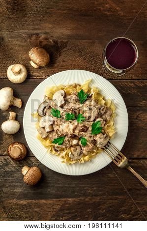 Mushroom beef stroganoff, served with typical egg pasta, with cremini and champignons, with glass of red wine, and fork, shot from above on dark rustic texture with place for text, vertical framing