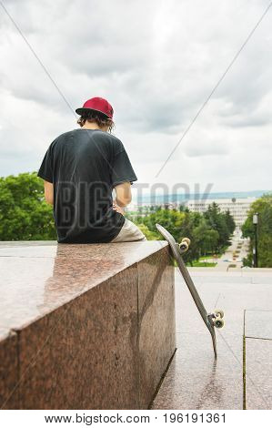 The long-haired skater in a cap and T-shirt sits with his back to the camera and thinks next to the skateboard against the background of the landscape landscape.
