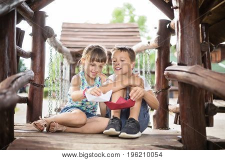 Two young caucasian children sitting next to each other with bent knees in a wooden house outdoors on sunny summer day playing with paper boats
