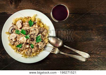 Mushroom beef stroganoff, served with the typical egg pasta, with cremini and champignons, with a glass of red wine, a fork, and a spoon, shot from above on a dark rustic texture with a place for text