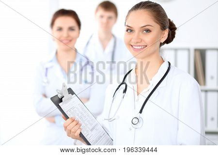Happy female doctor keeping medical clipboard while medical staff are at the background. Successful team at health care and medicine concept
