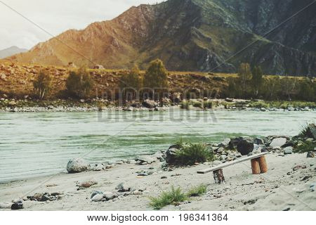 View of self-made wooden bench standing alone on sandy and rocky beach of fast Katun river; mountains hills and sparse trees in defocused background Altai Kuyus district Russia