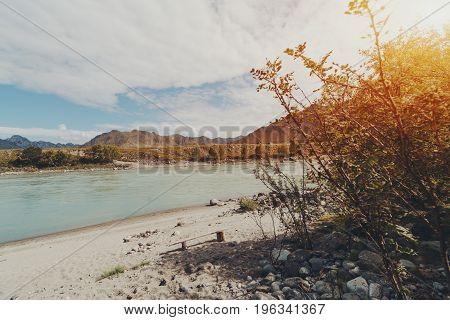 Beautiful morning wide angle view through bushes on wooden self-made bench lonely standing on sand beach of Katun river with stones mountains in distant Altai Kuyus district Russia