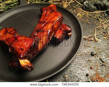 Fried oil ribs lie on a black frying pan. The frying pan stands on a dark wooden table among pieces of coal. Hay is lying around the frying pan.
