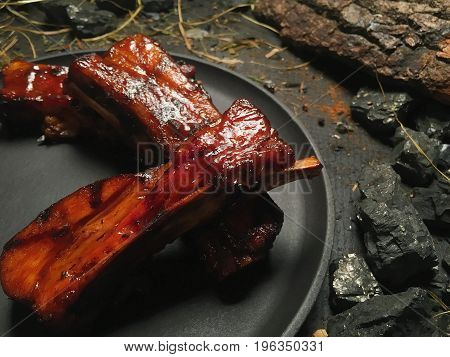 Fried ribs lie on top of each other. Close-up. Fried oil ribs lie on a black frying pan. The frying pan stands on a dark wooden table among pieces of coal. Hay is lying around the frying pan.