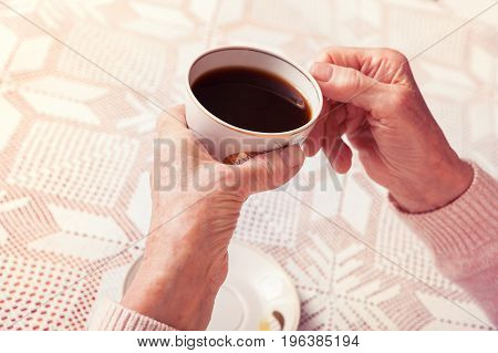 An elderly woman drinks tea at home. Senior woman holding cup of tea in their hands at table close-up