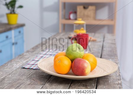 Close up view on fruit on a table at kitchens wooden table. Bright apples and oranges on front view with teaspot and red mug on background.