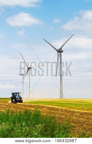 row of eolian wind turbine on a meadow with tractor ploughing the land in the background on a sunny day blue sky with clouds