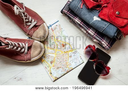 Travel accessories: hipster sneakers, denim shorts, plaid shirt, cowboy belt, hat, sungkasses, map and phone with headphones on wooden background. Selective focus. Traveling concept