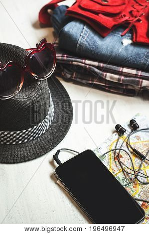 Travel accessories: hipster sneakers, denim shorts, plaid shirt, cowboy belt, hat, sungkasses, map and phone with headphones on wooden background. Selective focus. Traveling concept