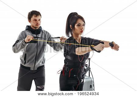 Electrical muscular stimulation fitness woman in full ems suit doing chest crossfit exercise with training cable. Trainer helps her. Studio photo isolated on white background.