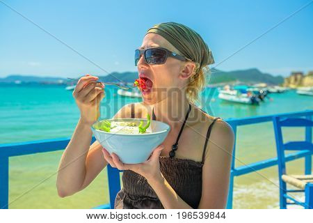 Happy woman eating healthy typical Greek salad at outdoor restaurant. Healthy Mediterranean diet concept. Methoni Beach on background in Greece. Woman enjoying healthy food in summer vacation