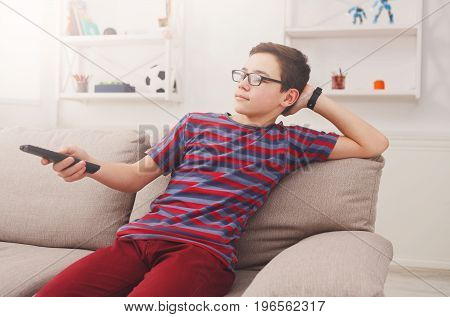 Teenager boy watching television, using TV remote while sitting on couch in living room at home