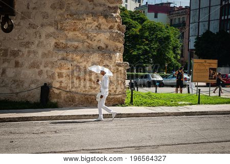 Havana, Cuba - June 30, 2016; member of Santeria Cuban religious sect dressed in traditional all white with umbrella contrasts with rough surrounds as he walks along city street past old stone museum building Havana.