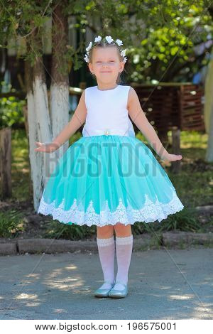 beautiful little girl in dress standing and posing over nature background child with a wreath of artificial flowers on her head look at the camera full length