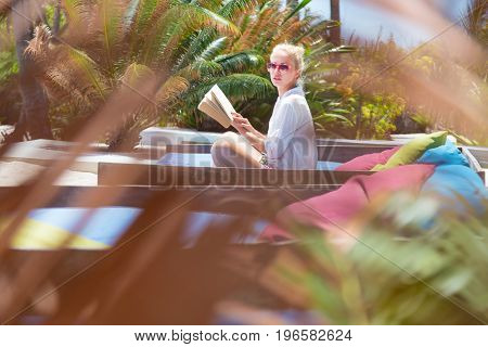 Fancy lady in white beach tunic reading book and relaxing by poolside of luxury spa resort set in lush tropical garden. Relaxation and wellbeing concept. Shot trough palm tree leaves.