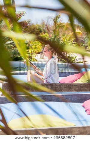 Fancy lady in white beach tunic reading book and relaxing by poolside of luxury spa resort set in lush tropical garden. Relaxation and wellbeing concept. Shot trough palm tree leaves.