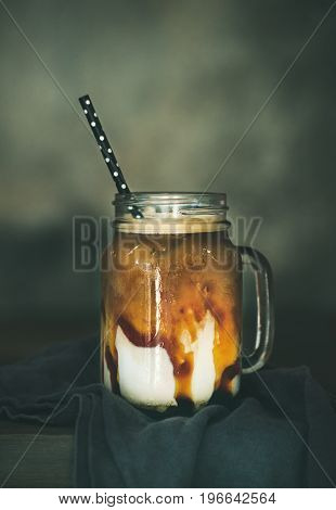 Iced caramel macciato coffee with milk in glass jar with straw on dark rustic wooden table, selective focus, copy space, vertical composition. Cold sweet refreshing summer drink concept