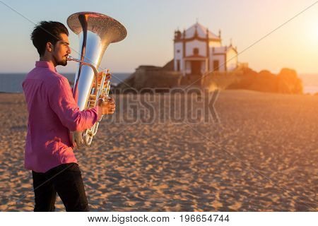 Young man playing the trumpet on the sea coast. Tuba instrument. In the background is the Chapel Senhor da Pedra at Miramar Beach, Atlantic ocean, Porto, Portugal.