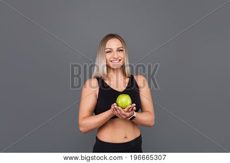 Blonde beefy woman wearing black sportswear holding apple with crossed hands standing against gray background.