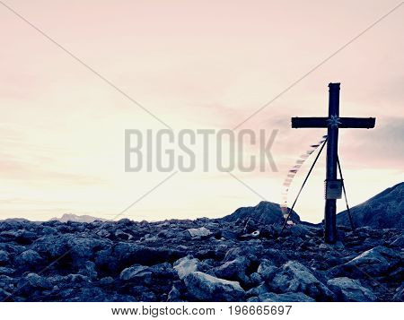 Modest Wooden Cross Raised On Rocky Alpine Mountain Summit . Sharp Rocky Peak. Gentle Clouds  In Blu