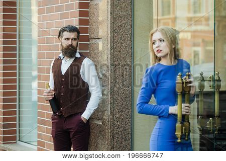 Man with bottle of wine looking at girl opening glass door. Sexy woman in blue dress and brutal hipster outdoors on brick wall. Alcohol and convive. Unhealthy lifestyle. Bad habits