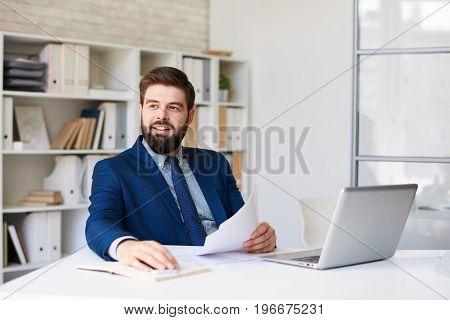 Portrait of successful  bearded businessman working at desk in light office looking away pensively and smiling
