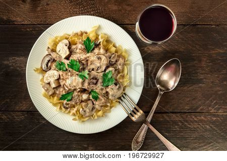 Mushroom beef stroganoff, served with the typical egg pasta, with cremini and champignons, with a glass of red wine, a fork, and a spoon, shot from above on a dark rustic texture with a place for text