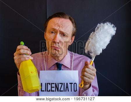 a sad man with the sign around his neck fired in italian language, holds the detergent and the duster / a man is sad because he was fired and has to deal with to do the cleaning work with the detergent and the duster