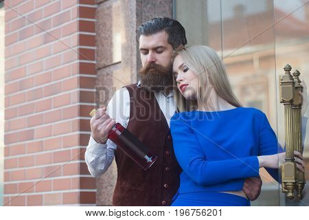 Couple in love looking at bottle of wine. Man or hipster hugging sexy woman or girl in blue dress outdoors on brick wall. Alcohol and convive. Unhealthy lifestyle. Bad habits