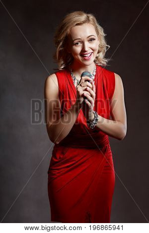 half length portrait of beautiful young woman in red dress holding microphone in her hand singing satanding next to color background