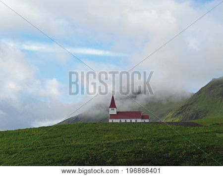 lonely red white church in vik in iceland green grass hills and low clouds