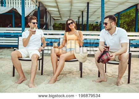 Lifestyle Young people enjoying summer vacation sunbathing drinking at beach bar. Two guys and a young girl sit on the terrace of the summer bar and enjoy the conversation