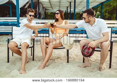 Lifestyle Young people enjoying summer vacation sunbathing drinking at beach bar. Two guys and a young girl sit on the terrace of the summer bar and enjoy the conversation