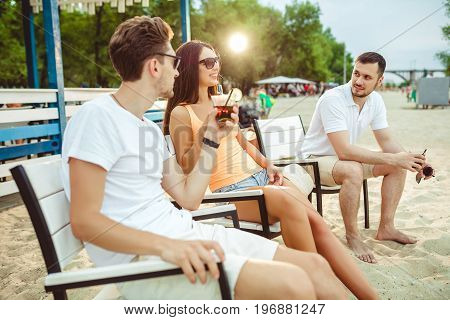 Lifestyle Young people enjoying summer vacation sunbathing drinking at beach bar. Two guys and a young girl sit on the terrace of the summer bar and enjoy the conversation