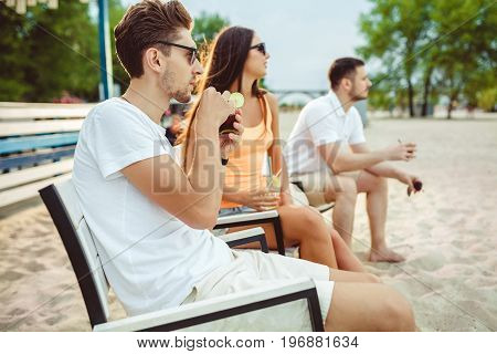 Lifestyle Young people enjoying summer vacation sunbathing drinking at beach bar. Two guys and a young girl sit on the terrace of the summer bar and enjoy the conversation