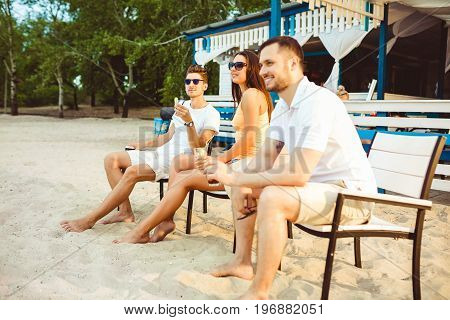 Lifestyle Young people enjoying summer vacation sunbathing drinking at beach bar. Two guys and a young girl sit on the terrace of the summer bar and enjoy the conversation