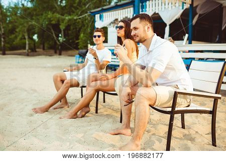 Lifestyle Young people enjoying summer vacation sunbathing drinking at beach bar. Two guys and a young girl sit on the terrace of the summer bar and enjoy the conversation