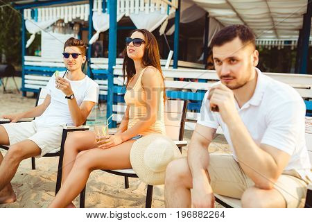 Lifestyle Young people enjoying summer vacation sunbathing drinking at beach bar. Two guys and a young girl sit on the terrace of the summer bar and enjoy the conversation