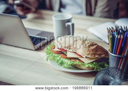 Close up view of working palce of freelancer during lunch. Wooden desk with laptop, cup of coffee and sandwich on it.