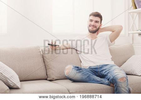 Young smiling man wathing tv at home. Happy guy pointing with remote control on tv-set, copy space
