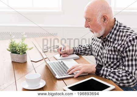 Mature man using laptop and writing in notepad at home desk. Education, recipe concept