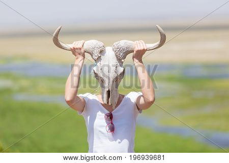 Woman holding a white wildebeest skull wearing it like a mask in nature on african wildlife safari, Amboseli national park, Kenya.