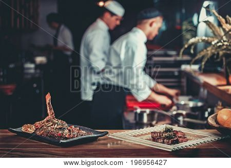 Food concept. Ready grilled pork ribs dish and beef steak with herbs. Ready to serve. Ready to eat. Two chefs working in the background interior of modern professional restaurant kitchen