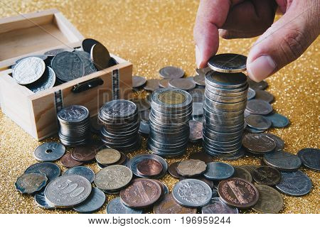 Hand putting a coin on stack of coins. Golden glittering background pile of coins in small wooden chest with coins from various countries scattering around.