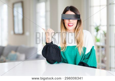 Young beautiful woman blindfold eyes with black censor band at home smiling with happy face looking and pointing to the side with thumb up.