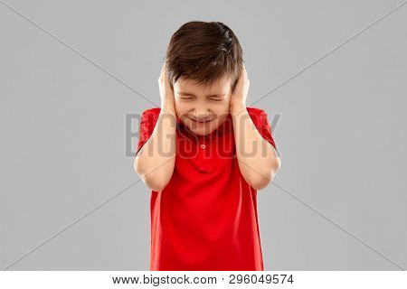 noise, stress and people concept - stressed little boy in red polo t-shirt closing ears by hands over grey background