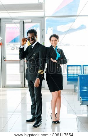 African American Pilot In Sunglasses And Stewardess With Crossed Arms Standing Together In Airport