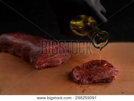 Hand in black glove pouring oil on beef steak with spices on a wooden board with a black background
